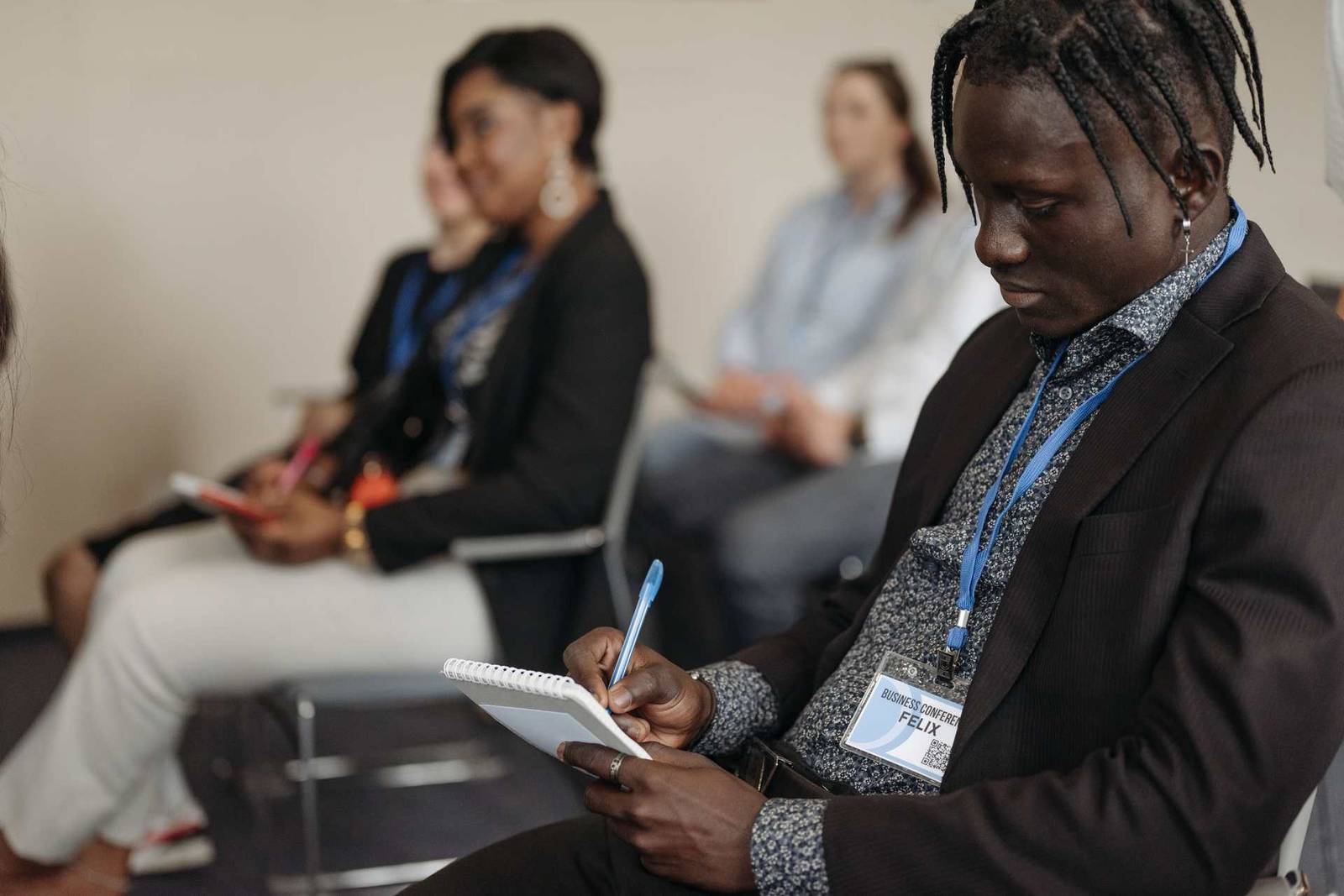 A conference attendee writes by hand in a spiral notebook, ID badge visible, seated among other attendees