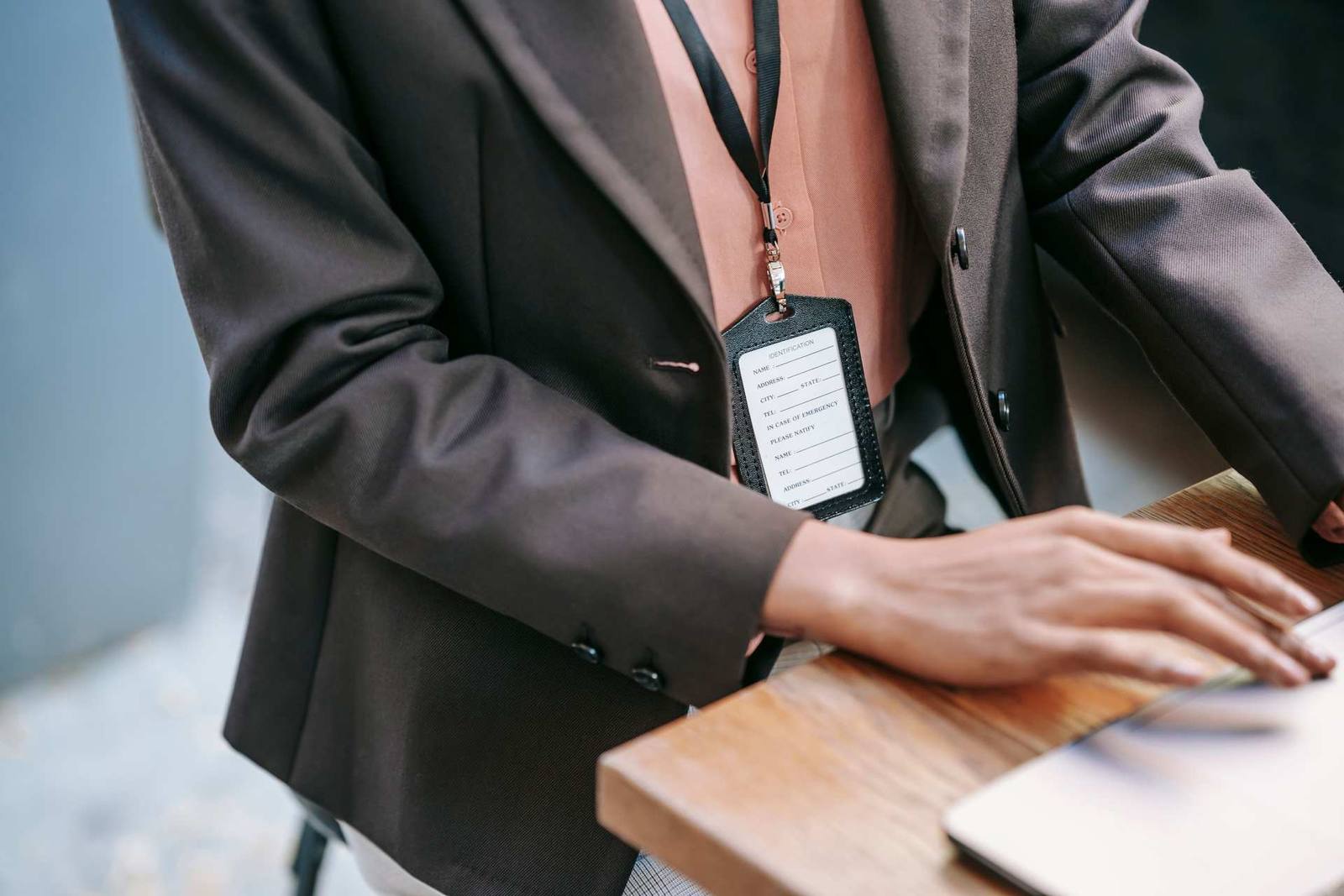 Close-up of a badge and lanyard against a blazer, a hand resting on a laptop at a wooden table