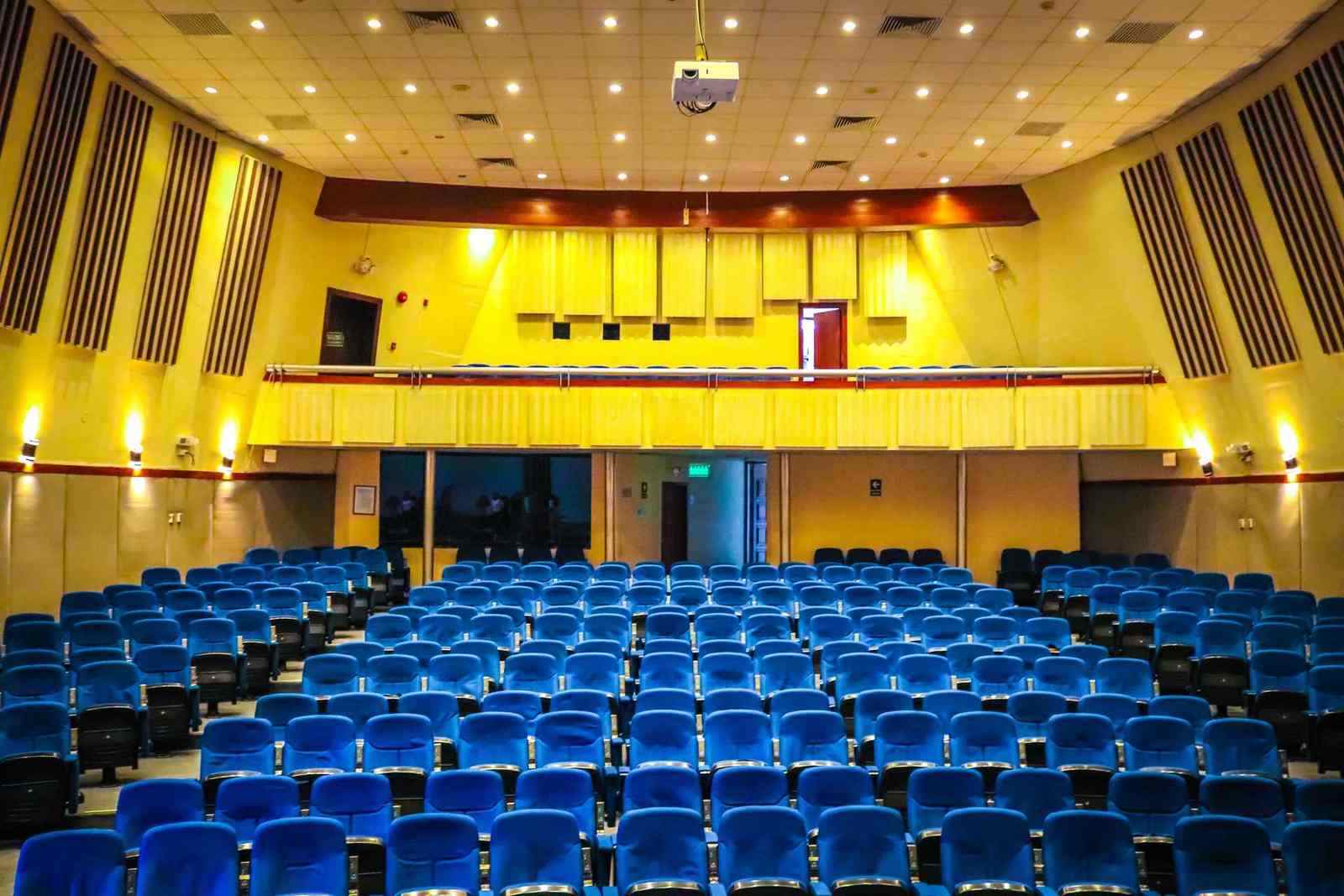 An empty auditorium with rows of blue upholstered seats, no people, facing a stage