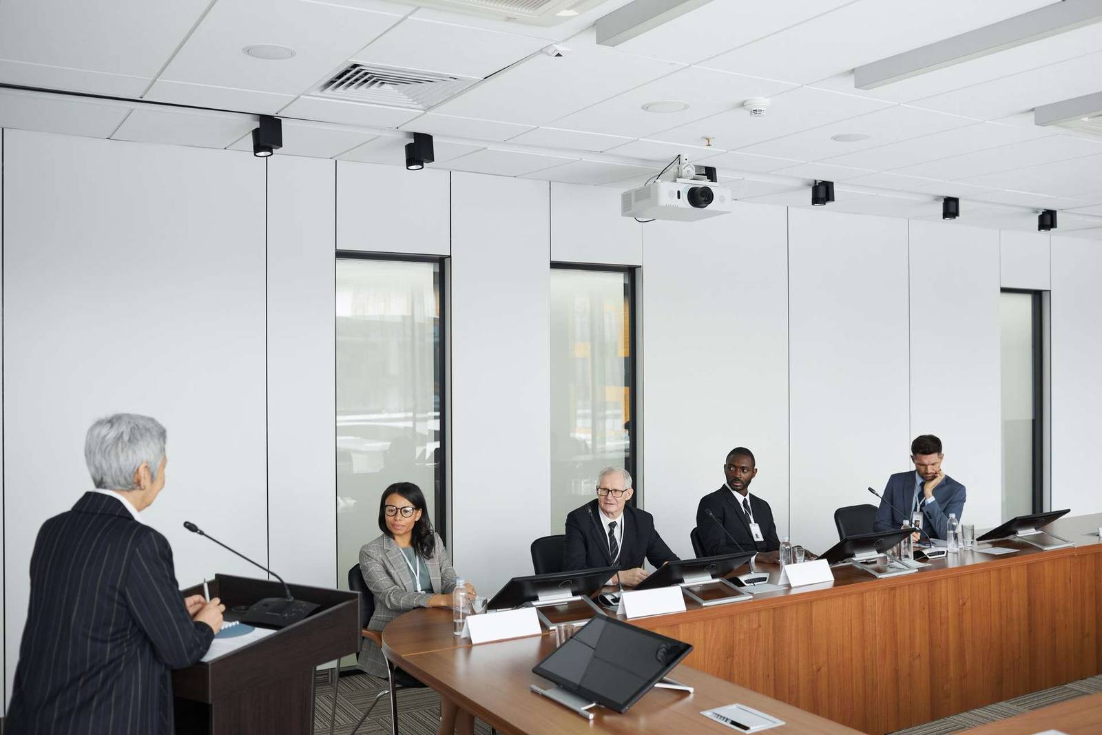 A committee chair addresses four seated colleagues at a formal meeting table, each with an individual monitor, in a wide conference room