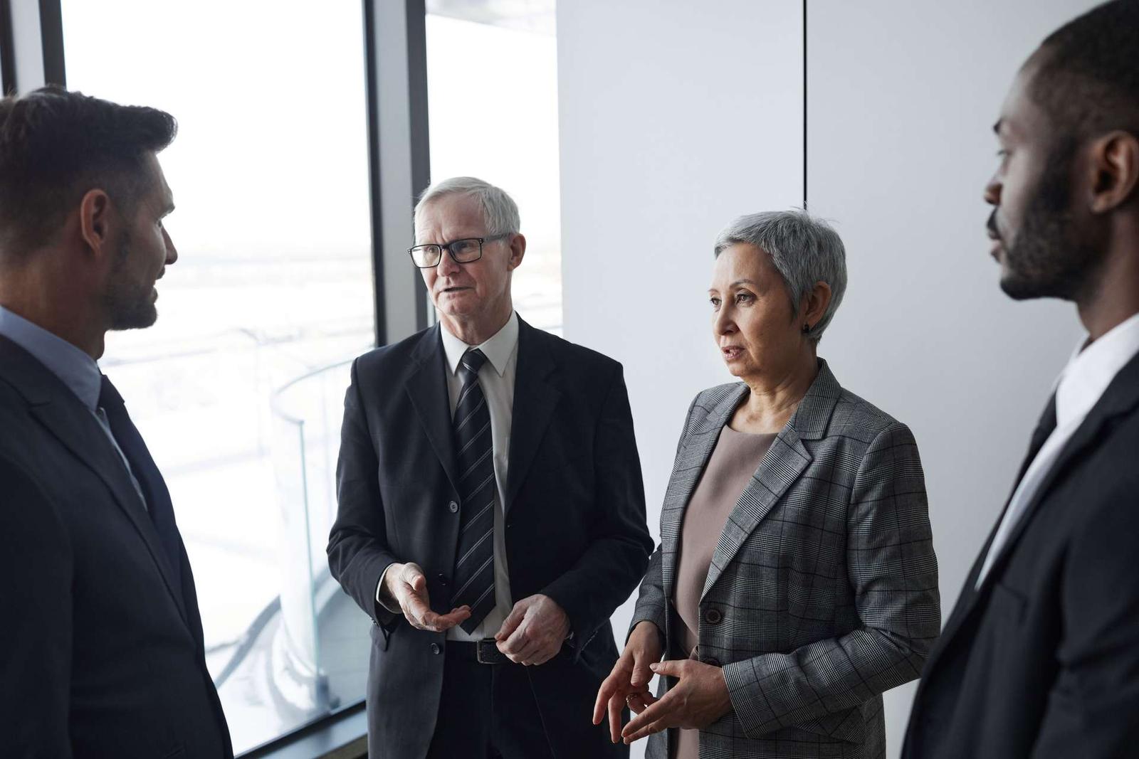 Four business-attired attendees stand in animated conversation near a window in a bright, modern venue