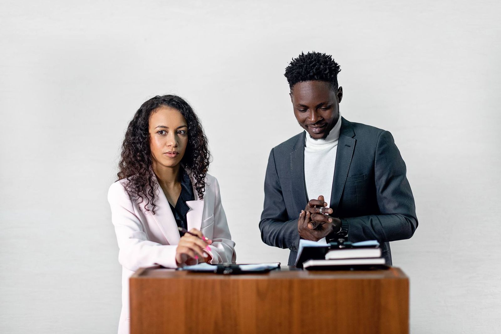 Two colleagues in business attire stand together at a wooden lectern, reviewing notebooks side by side against a plain light backdrop