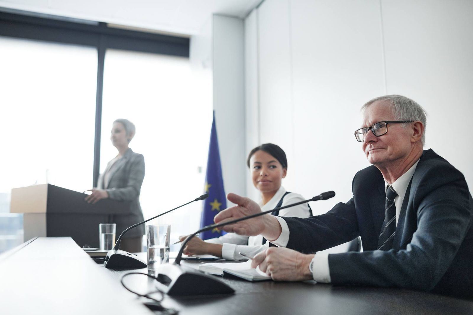An EU flag stands behind a panel of three speakers at microphones in a bright, formal conference room