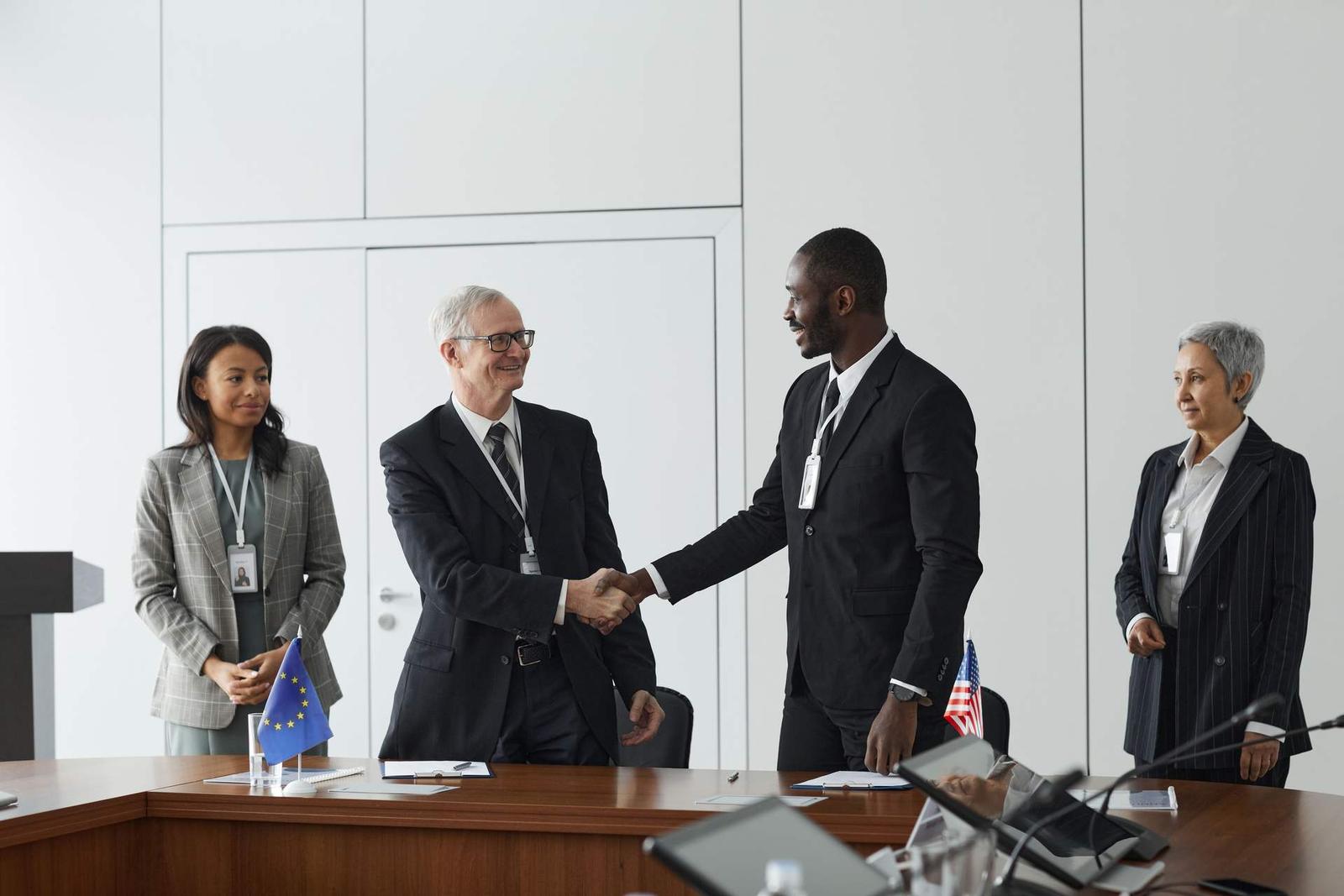 Four colleagues in business attire shake hands across a table bearing EU and US flags, in a bright formal room