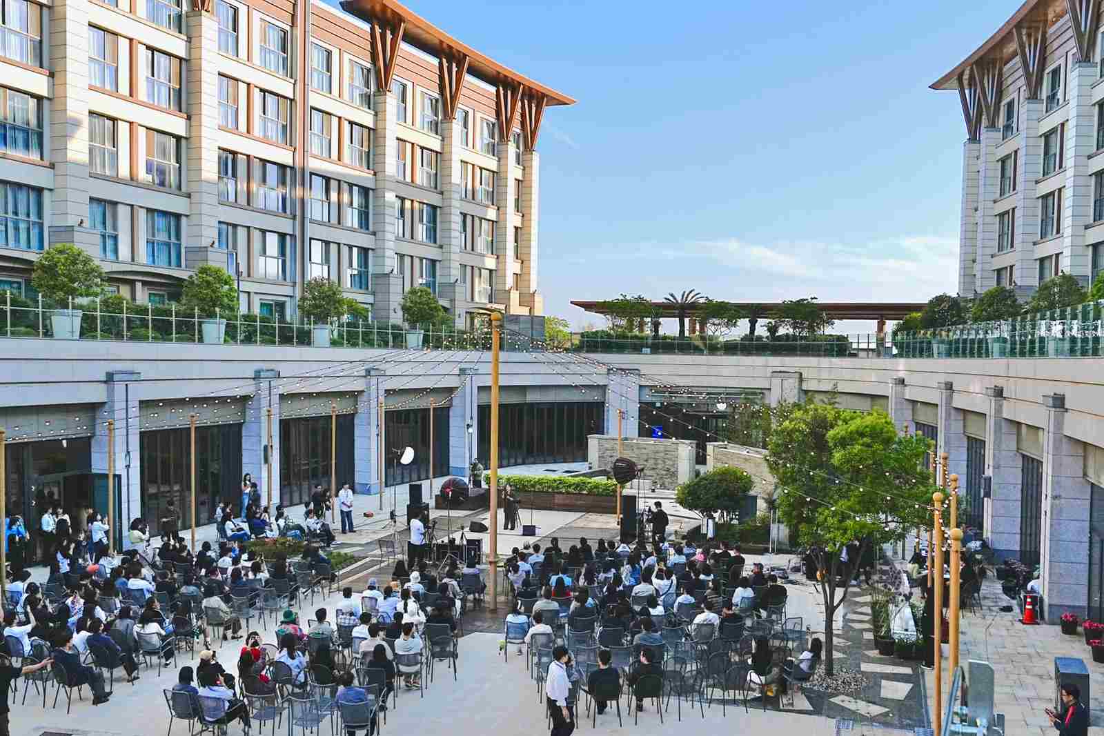 Rows of attendees at an outdoor evening reception seated facing a small lit stage, string lights strung overhead between two hotel-style buildings at dusk