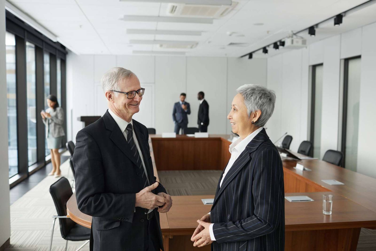 Two senior delegates chat and smile in profile beside an empty U-shaped hearing-room table before the room fills, floor-to-ceiling windows and two colleagues in soft focus behind them