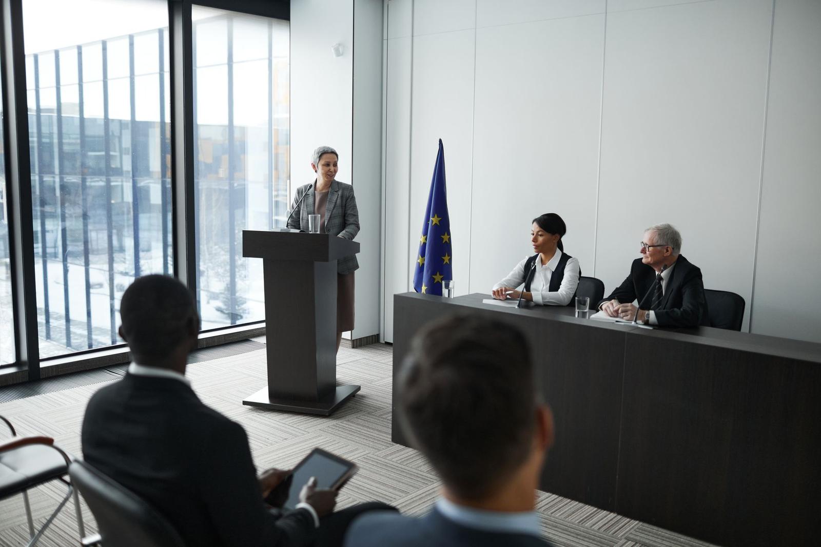 A speaker addresses a small panel seated beside a European Union flag in a formal conference room