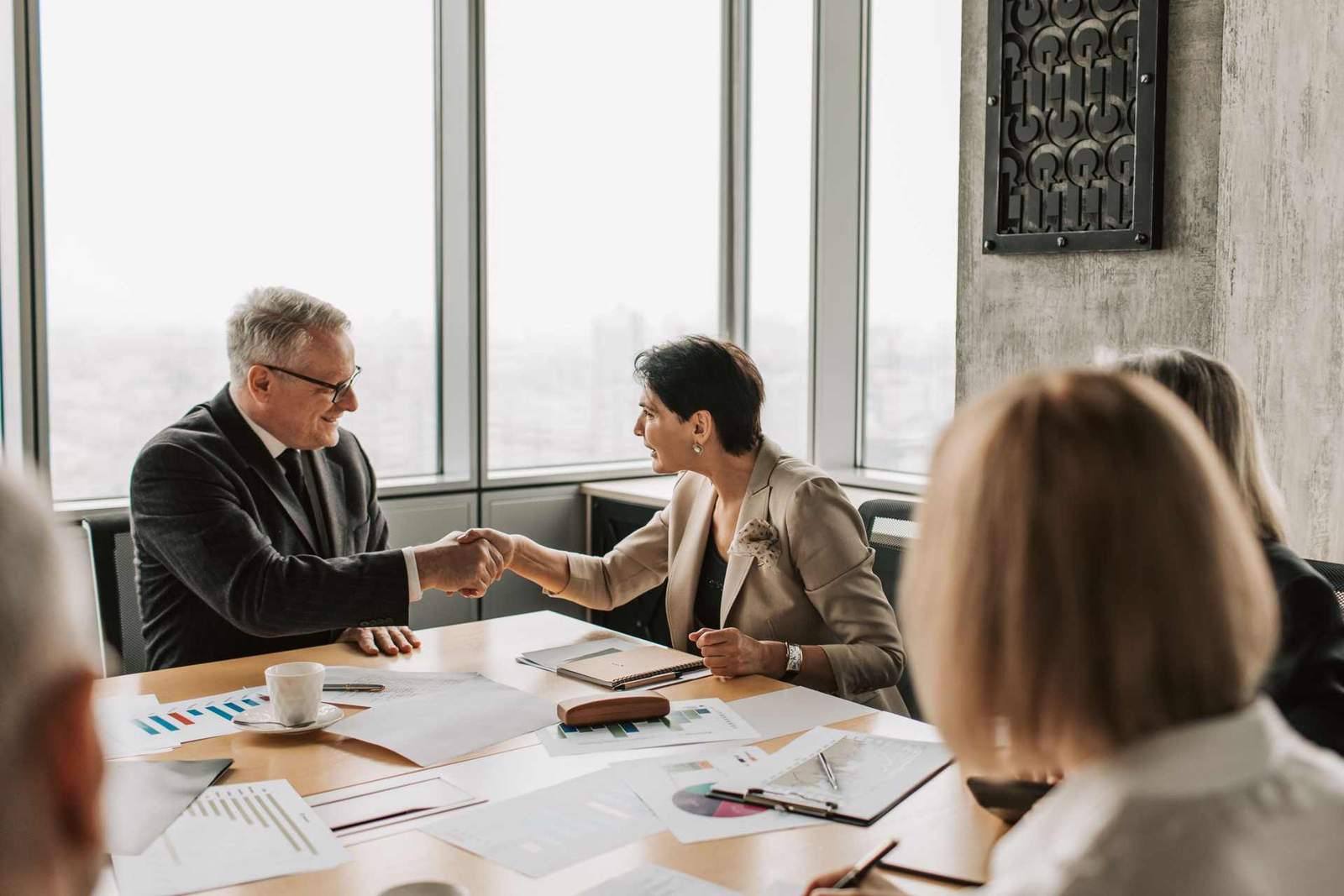 An older man and a woman in business attire shake hands across a table covered in printed charts, in a bright high-rise meeting room with colleagues visible nearby