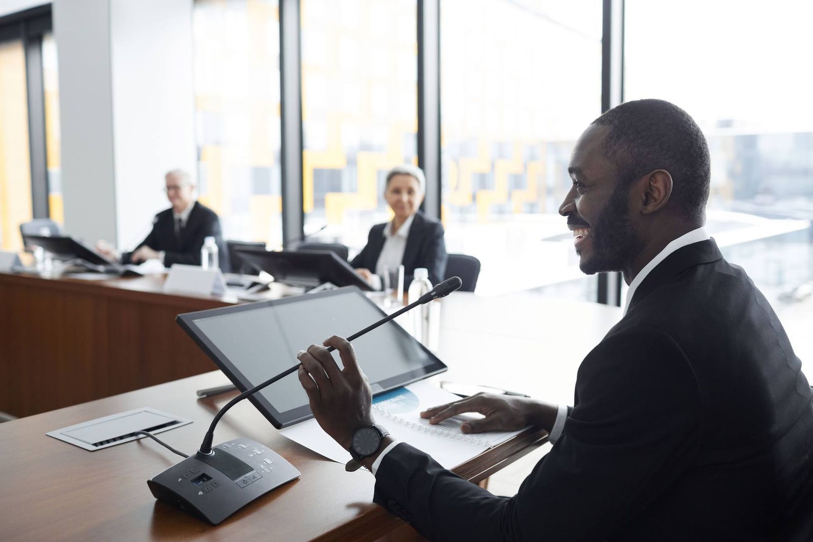 A delegate speaks into a desk microphone at a formal committee-style table, two colleagues visible at their own desks in soft focus with bright windows behind