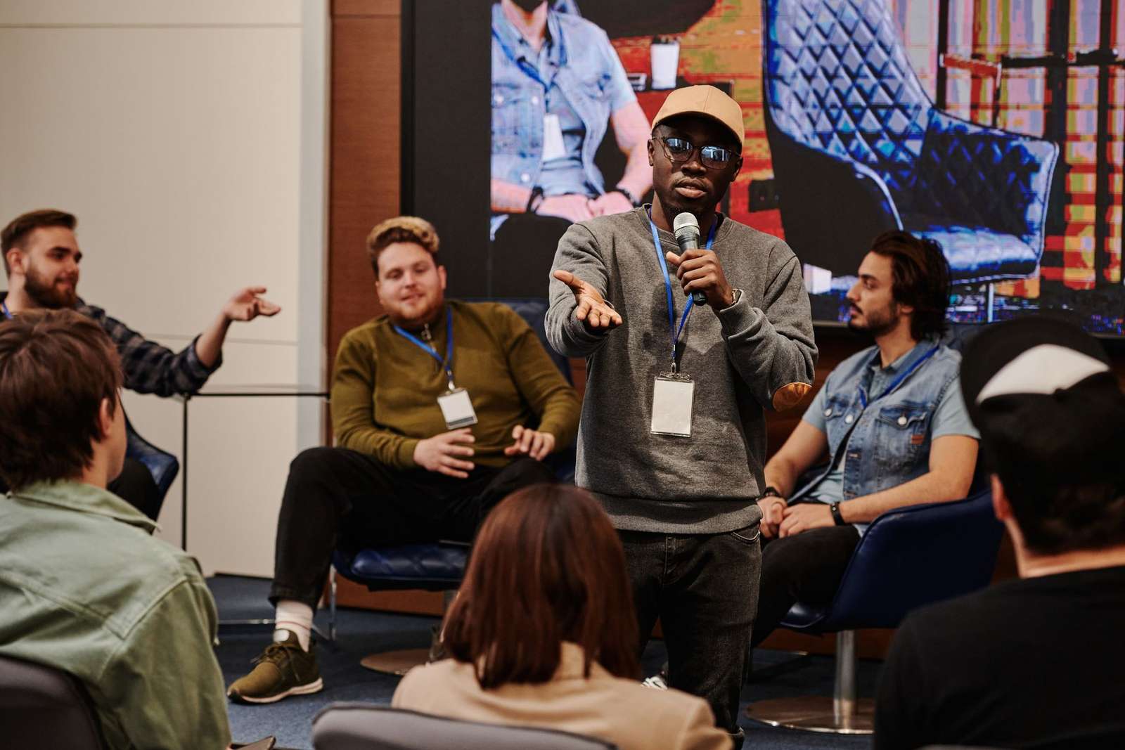 One person stands holding a microphone and gesturing mid-answer during a small panel discussion, seated colleagues and other attendees around them
