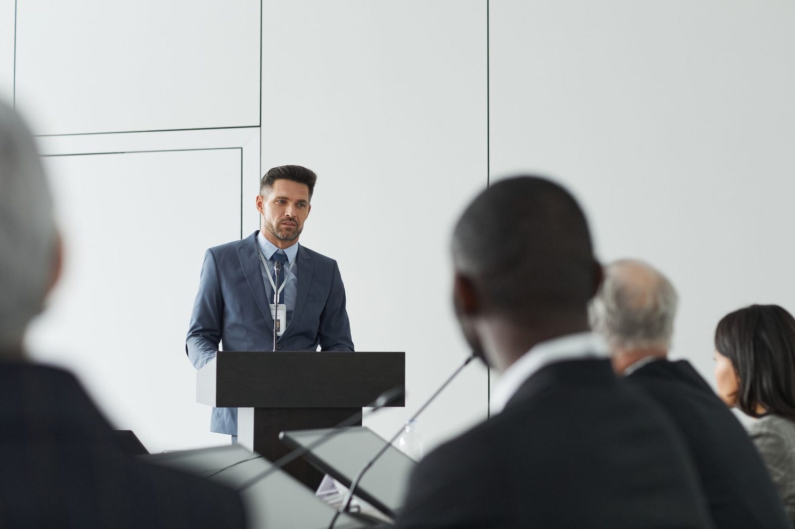 A speaker in a suit addresses the room from a lectern, seen past blurred audience members in the foreground
