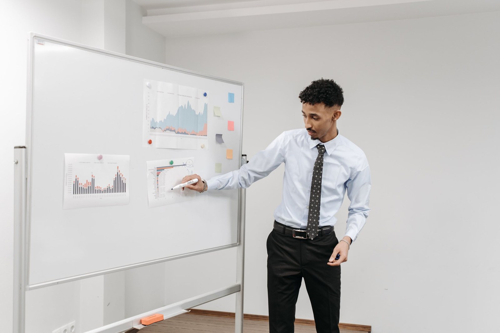 A presenter in a dress shirt and tie points to a printed chart pinned to a whiteboard covered in data printouts and sticky notes