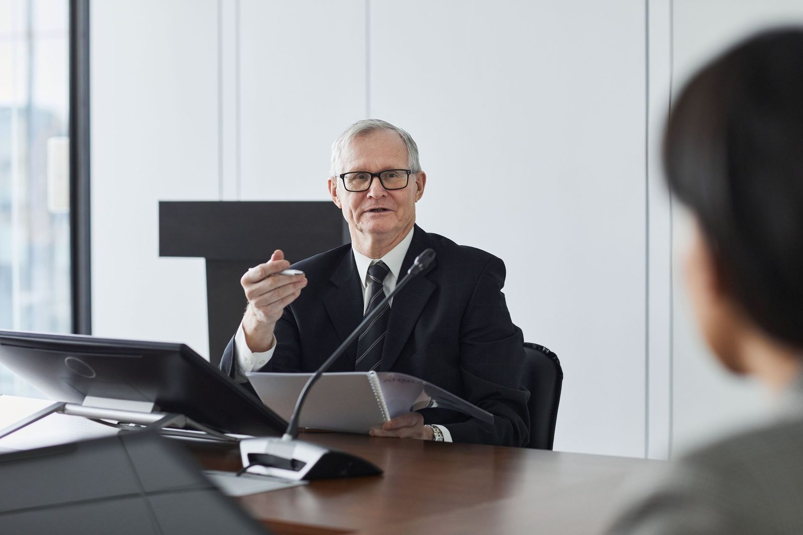 An older speaker in glasses gestures with a pen while speaking at a conference table, a monitor and spiral notebook in front of him, a colleague listening across the table