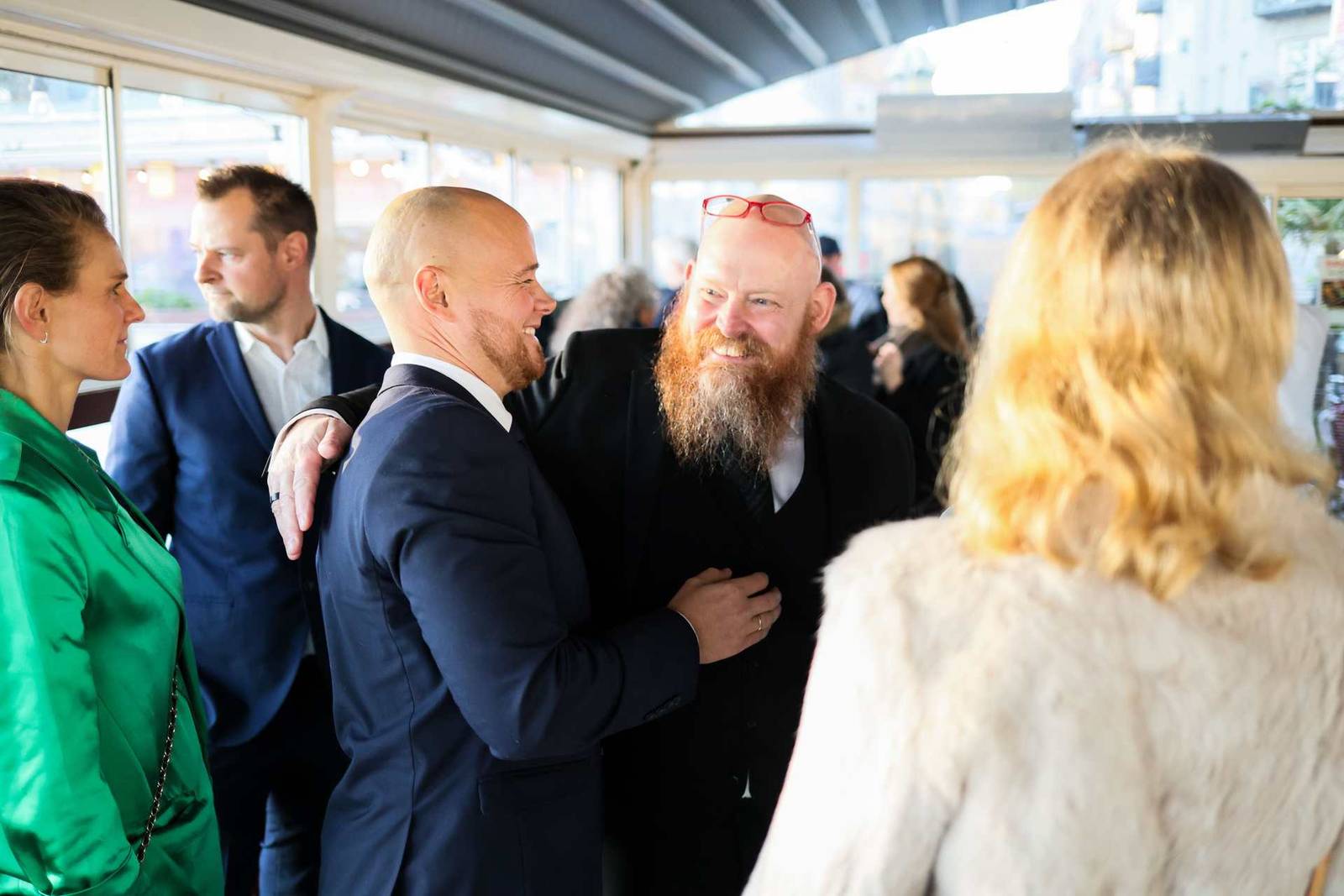 Two men embrace warmly in greeting amid a small group of colleagues, in a bright glass-walled reception space