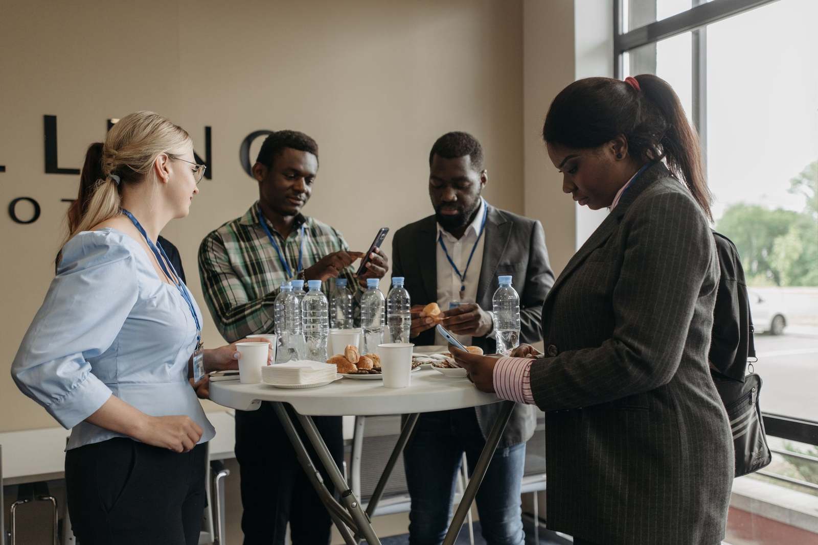 Four conference attendees standing around a break-table with coffee and pastries, one showing something on a phone to another