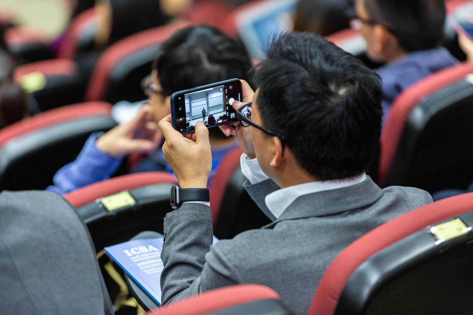 An attendee, seen from behind, photographing a speaker on stage with a phone in a packed auditorium