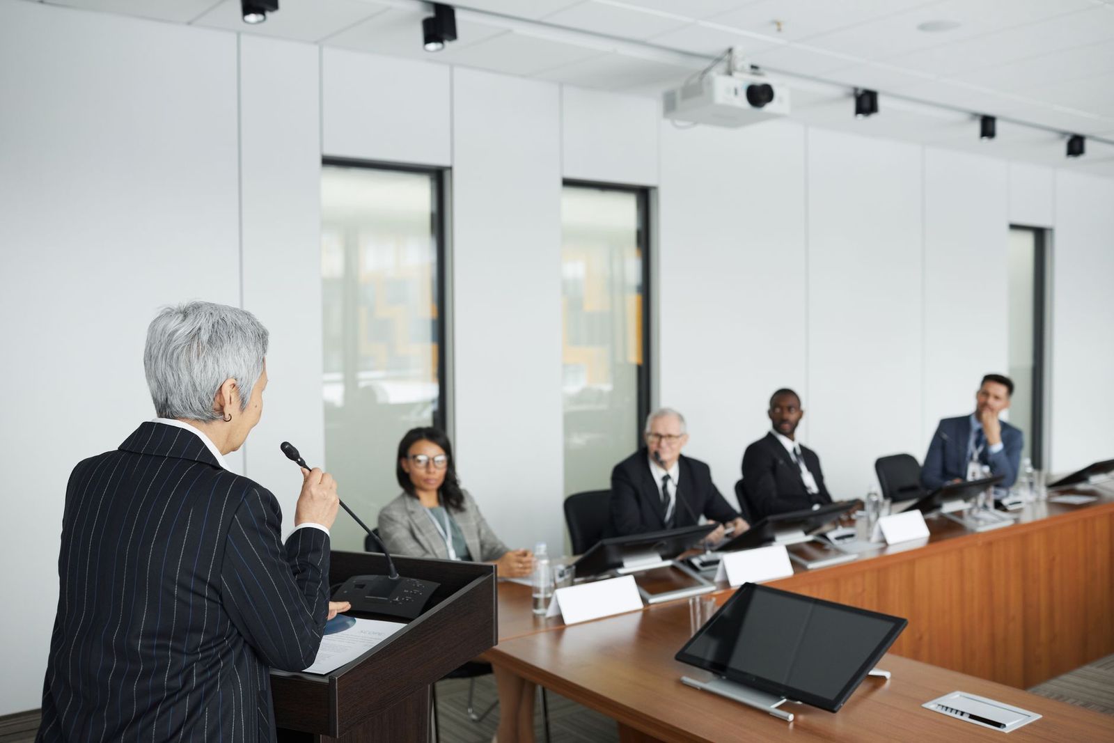 A speaker addresses a seated panel from a podium microphone in a hearing room