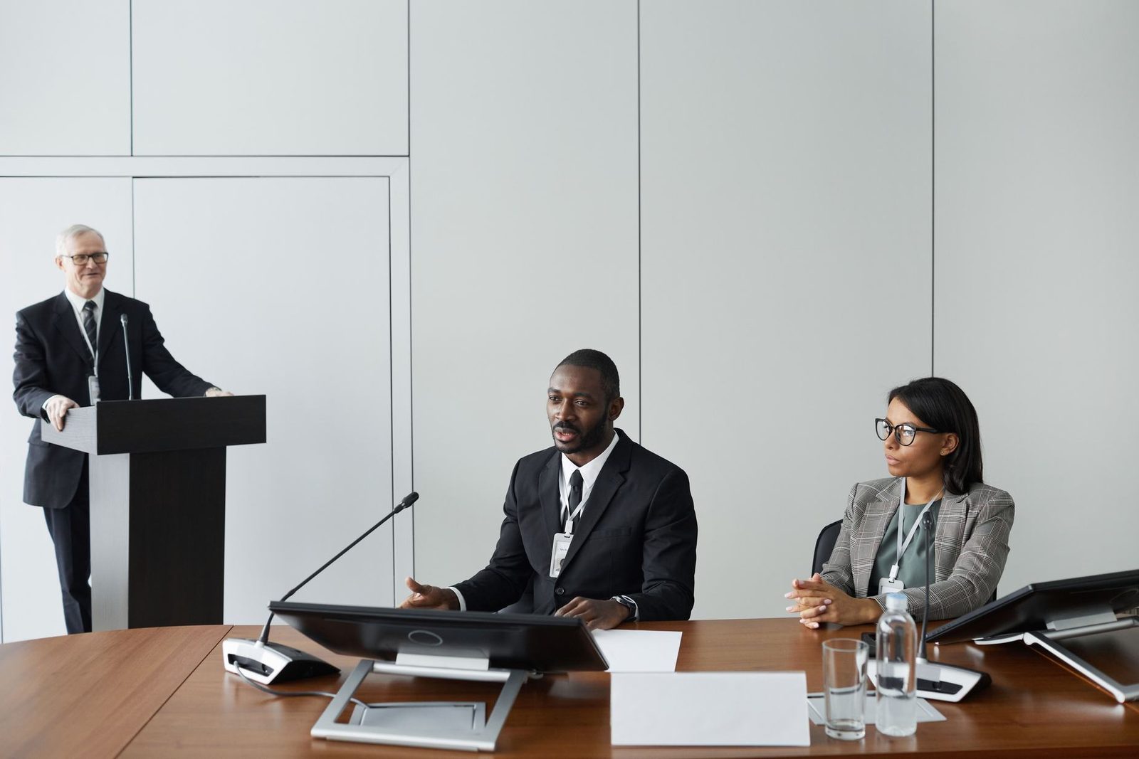 A seated panelist in a suit gestures while speaking at a conference table beside a colleague in a tweed blazer, with another presenter at a podium in the background