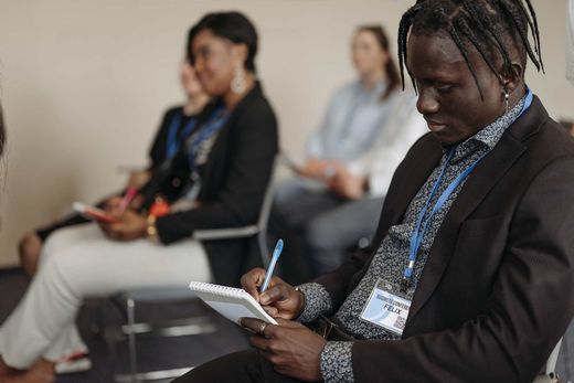 A conference attendee writes by hand in a spiral notebook, ID badge visible, seated among other attendees