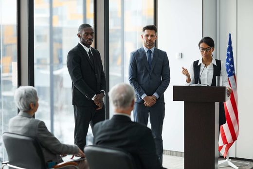 Three officials in dark suits stand before a lectern bearing a U.S. flag as a colleague speaks into a microphone, seated attendees in the foreground
