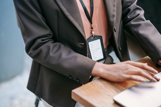 Close-up of a badge and lanyard against a blazer, a hand resting on a laptop at a wooden table