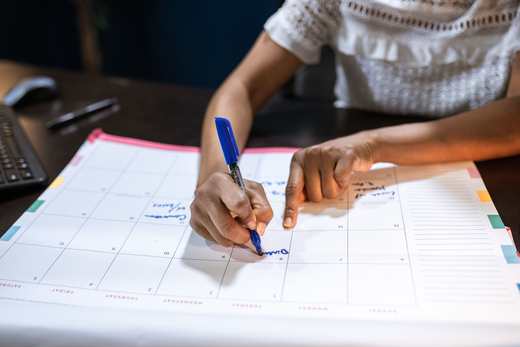 A hand marks dates on a large desk calendar with a blue pen