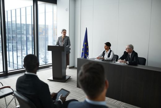 A speaker addresses a small panel seated beside a European Union flag in a formal conference room