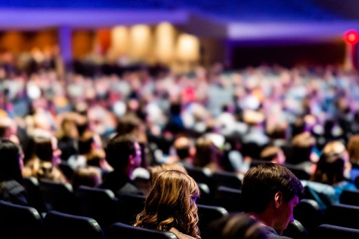 Attendees seated in a packed conference auditorium, seen from behind as a session begins