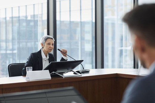 A panelist mid-answer at a desk microphone, addressing someone across the table in a formal meeting room