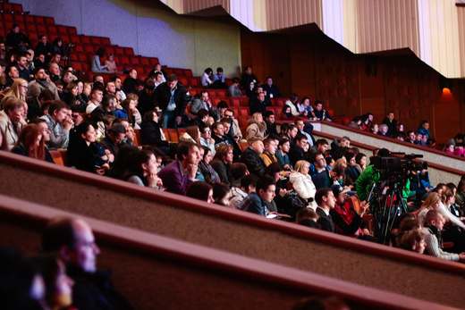 Rows of attendees fill a packed conference auditorium as a camera operator films from the aisle