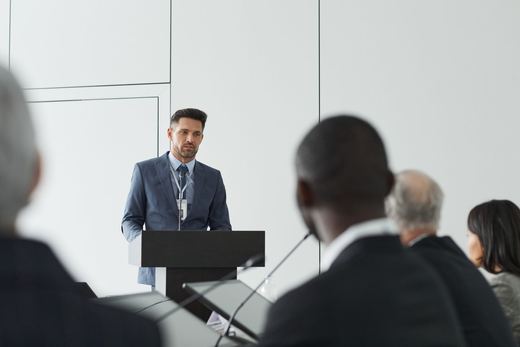 A speaker in a suit addresses the room from a lectern, seen past blurred audience members in the foreground