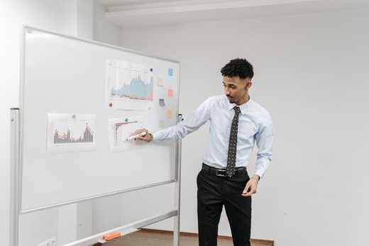 A presenter in a dress shirt and tie points to a printed chart pinned to a whiteboard covered in data printouts and sticky notes