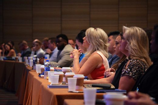 Rows of business-attired attendees seated at a linen-draped table, coffee cups and notebooks in front of them, listening to an unseen speaker in a ballroom