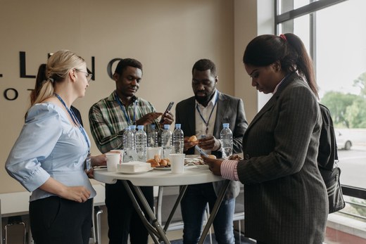 Four conference attendees standing around a break-table with coffee and pastries, one showing something on a phone to another