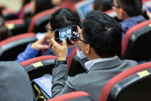 An attendee, seen from behind, photographing a speaker on stage with a phone in a packed auditorium
