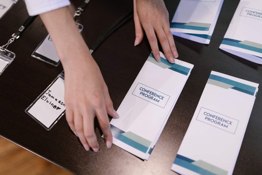Hands lay out conference programs, lanyards and a handwritten name badge at a registration table