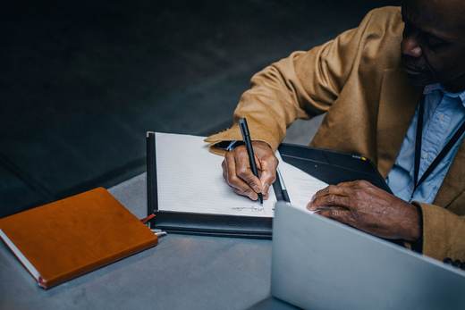 A man in a tan blazer writes notes in a leather folio beside an open laptop