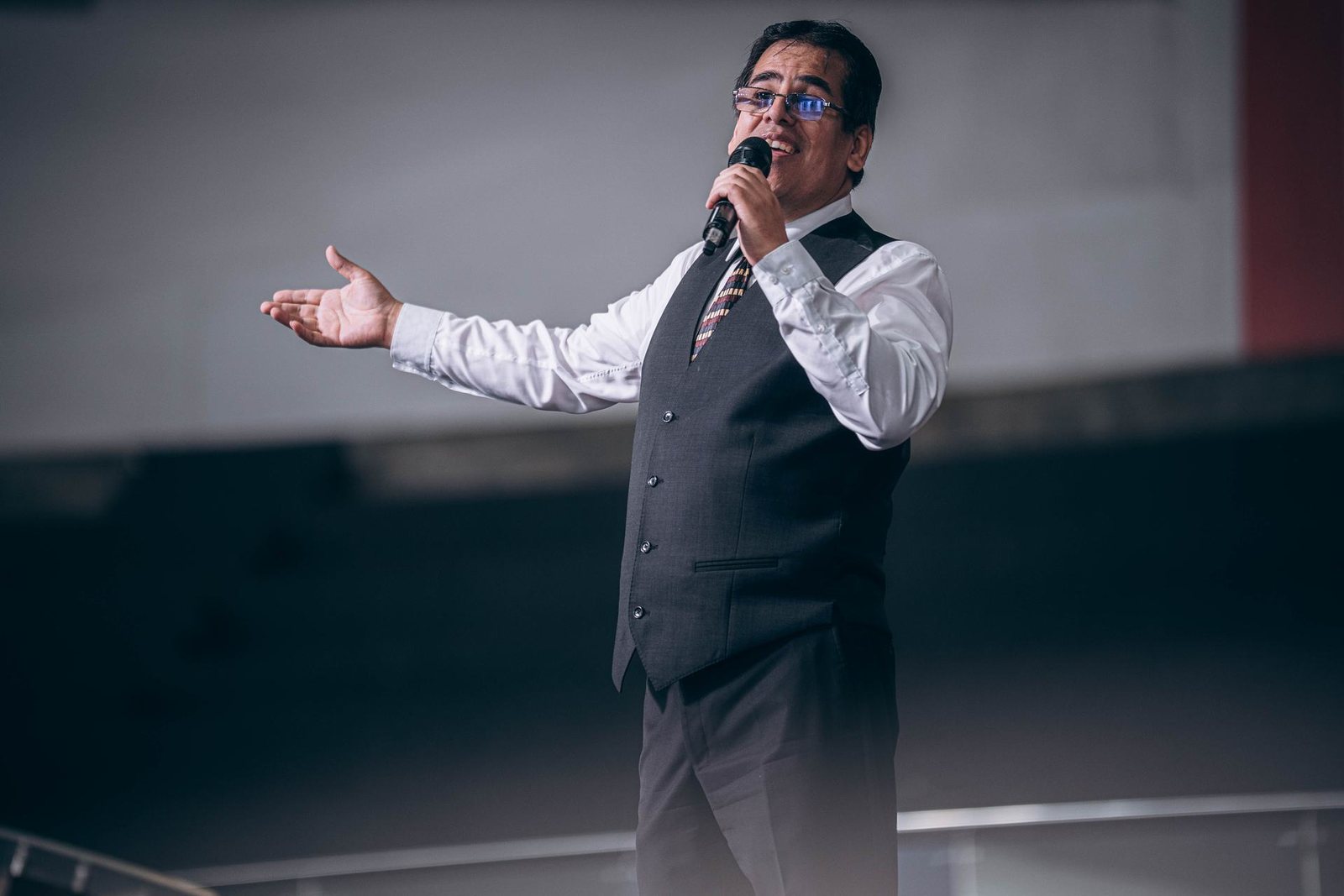 A speaker in a waistcoat and tie gestures with one arm outstretched while holding a microphone, on a dim stage