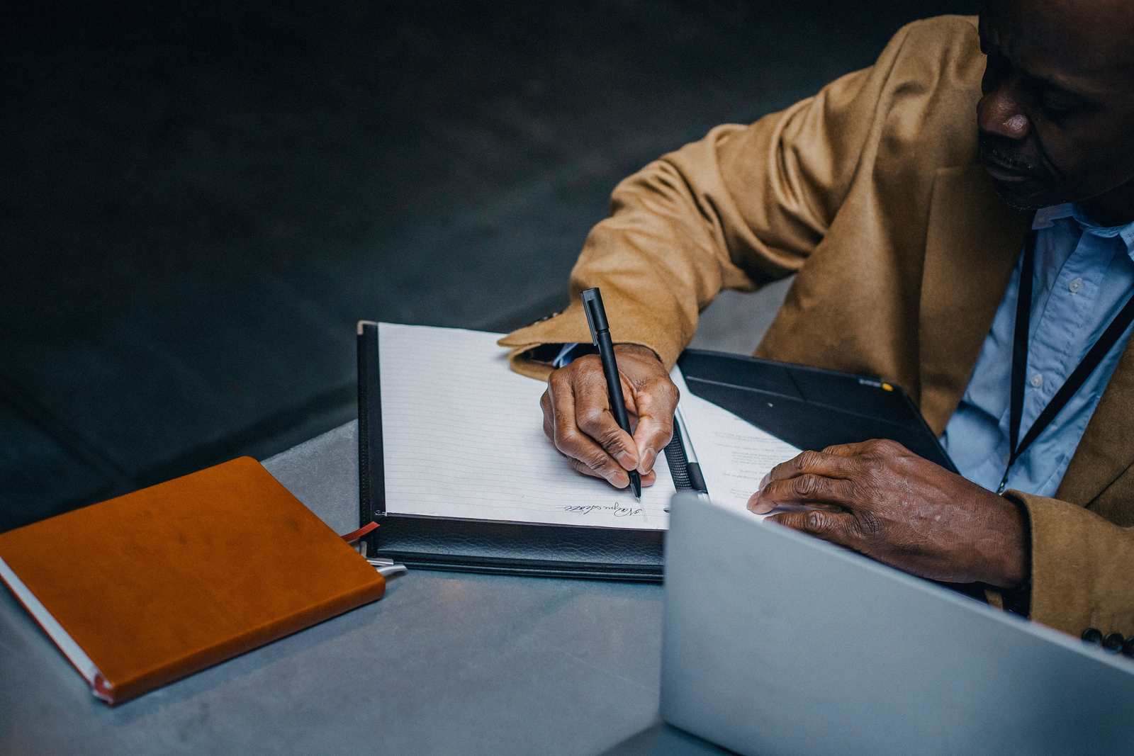 A man in a tan blazer writes notes in a leather folio beside an open laptop