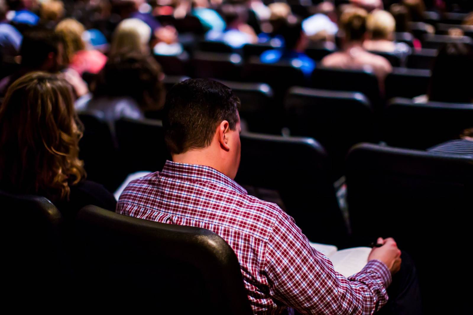 An attendee, seen from behind, takes handwritten notes in a dim, packed lecture hall as a session runs