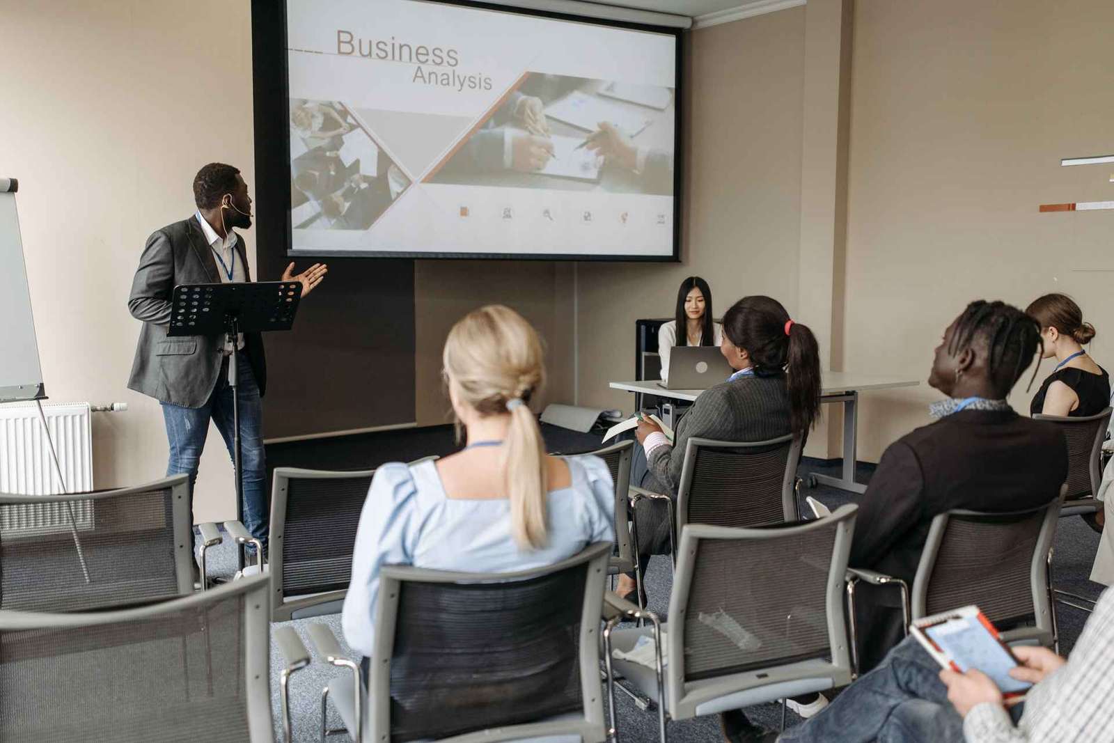 A presenter gestures while addressing a small seated group in a meeting room, a projection screen reading 