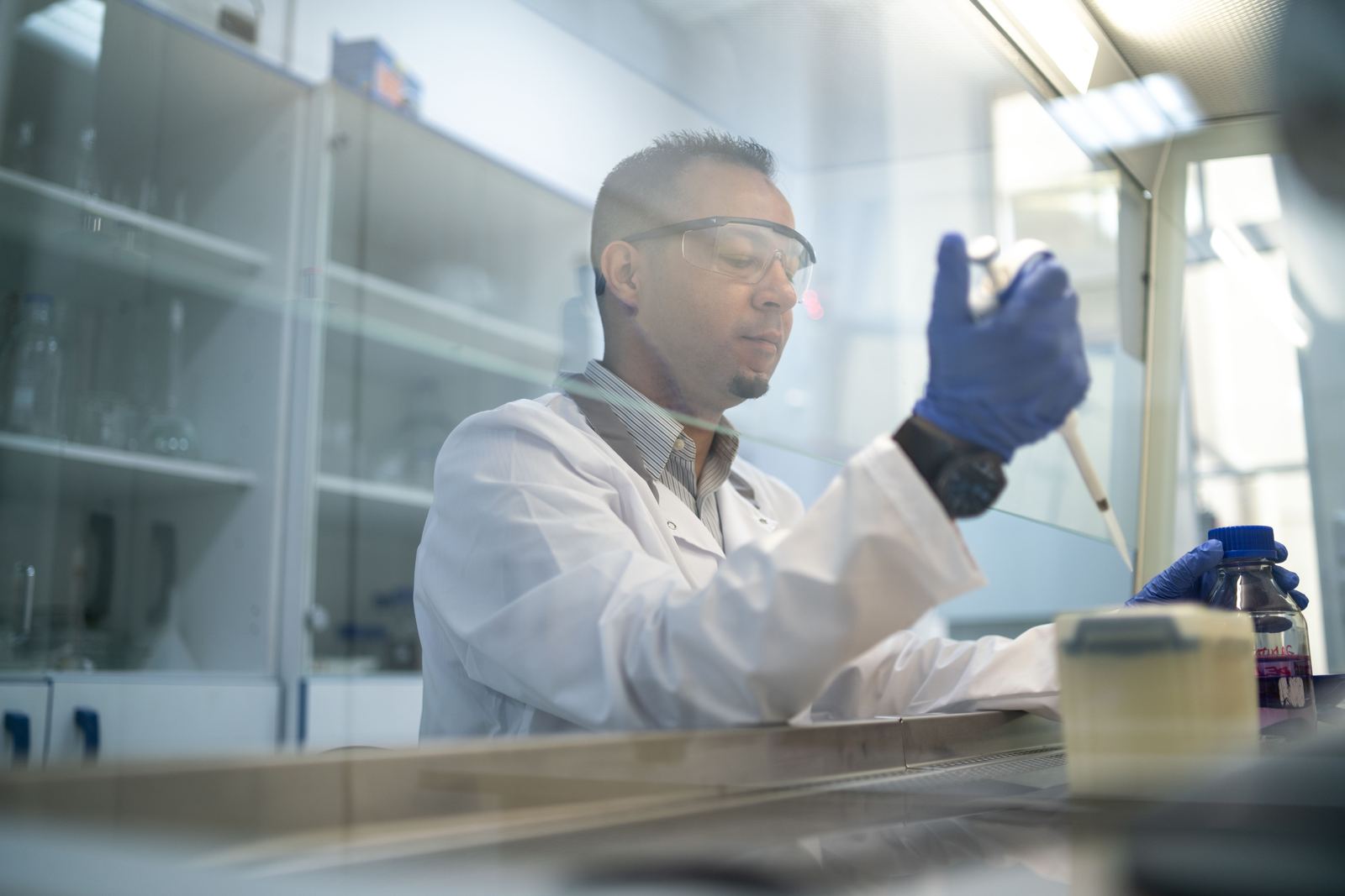 A scientist in safety glasses and blue gloves pipetting inside a biosafety cabinet