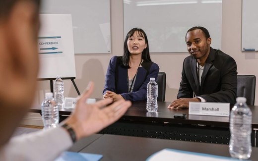 Two panelists seated at a conference table answering a question, with a CONFERENCE sign visible behind them