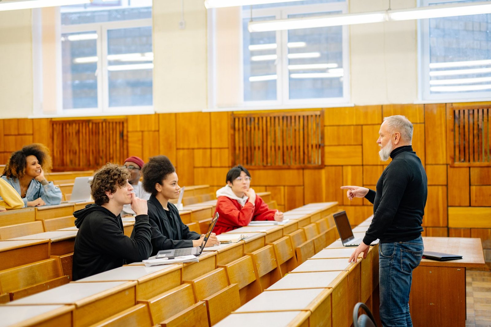 An instructor gestures while addressing students seated at desks in a tiered lecture hall