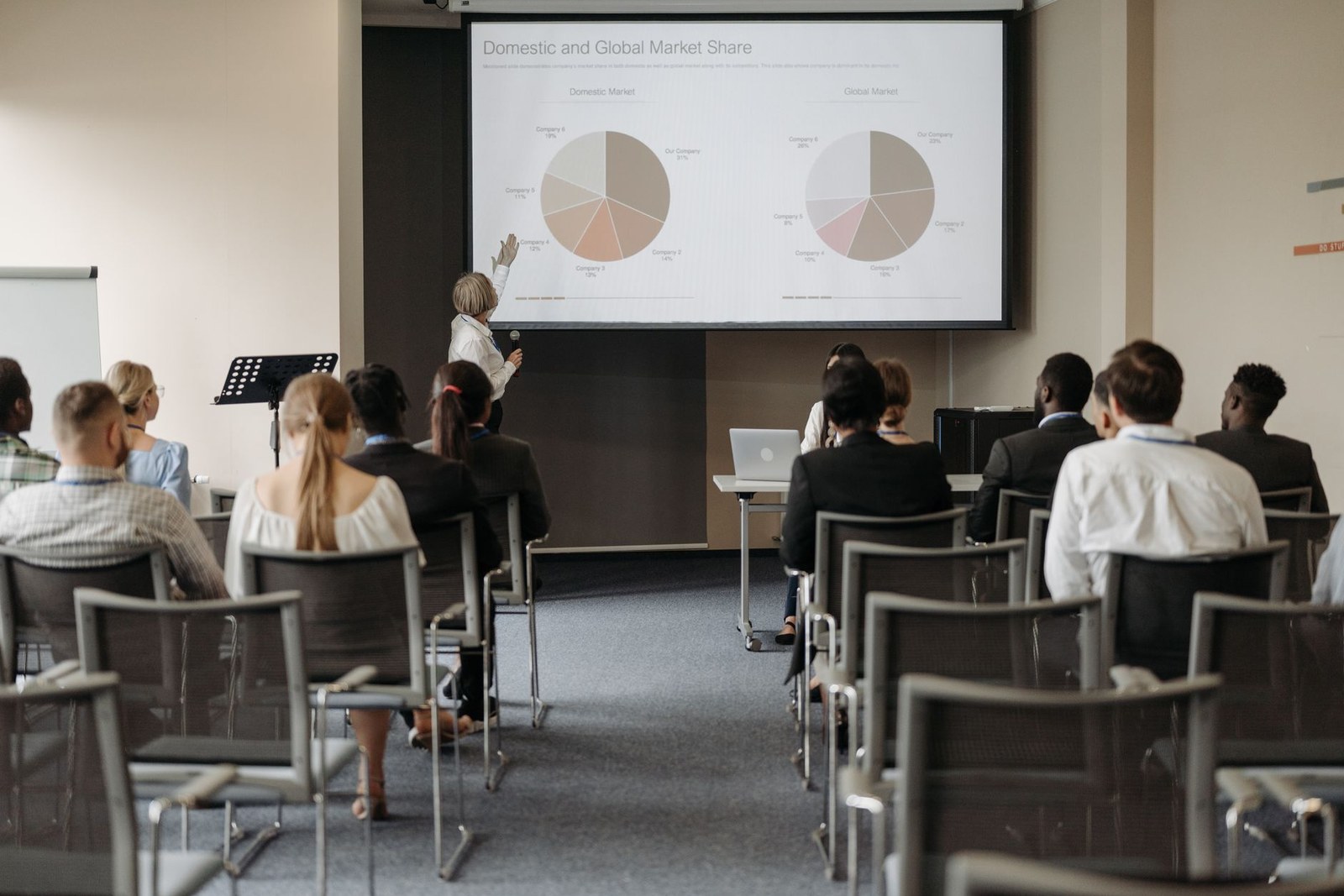 A presenter gestures toward a data-filled slide in front of a seated conference audience