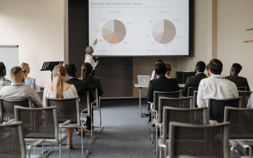 A presenter gestures toward a data-filled slide in front of a seated conference audience