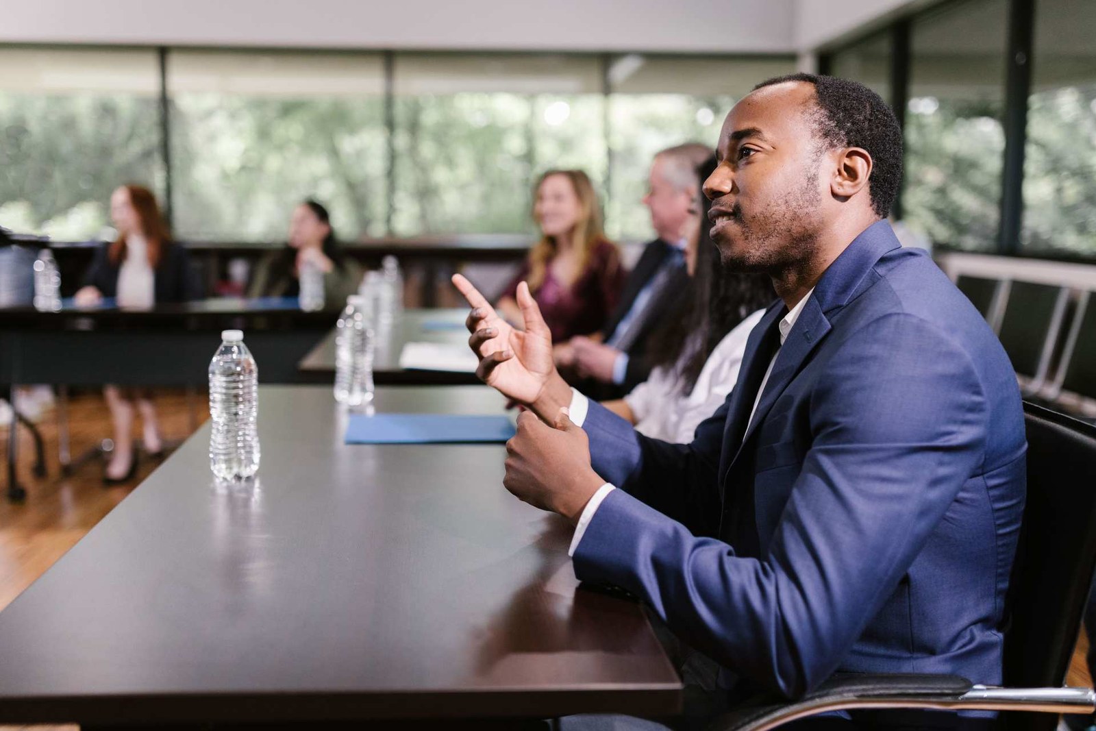 A man in a navy suit gestures while speaking at a conference table, with other attendees seated nearby