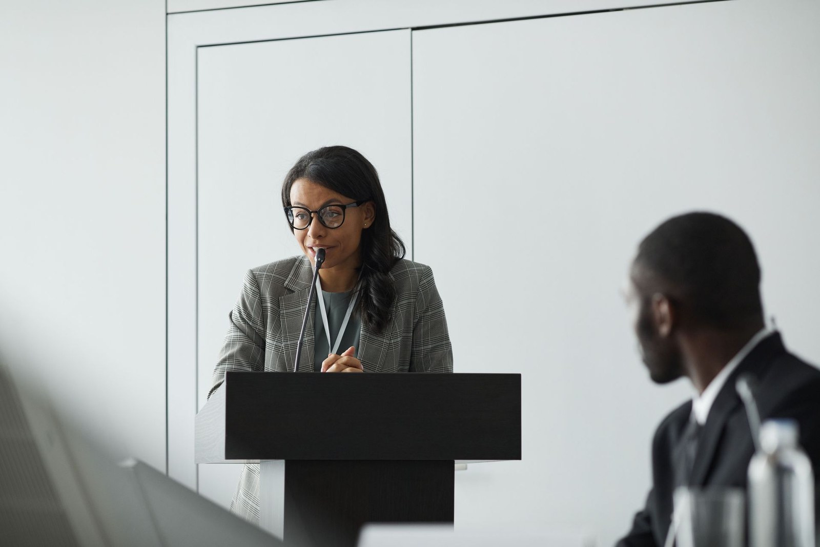 A speaker at a podium, wearing a conference lanyard, addresses the room during a formal panel proceeding