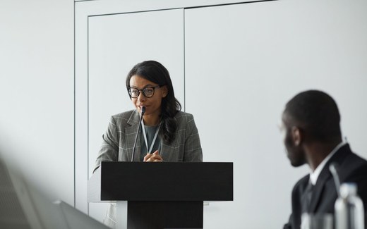 A speaker at a podium, wearing a conference lanyard, addresses the room during a formal panel proceeding