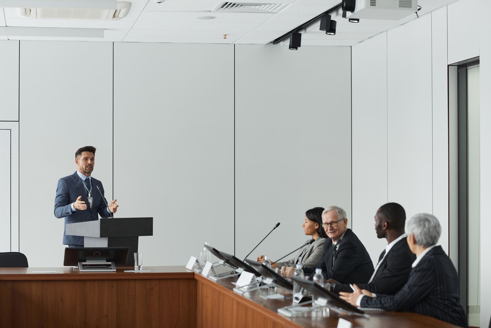 A speaker at a lectern gestures while addressing a panel of four seated colleagues in a conference room